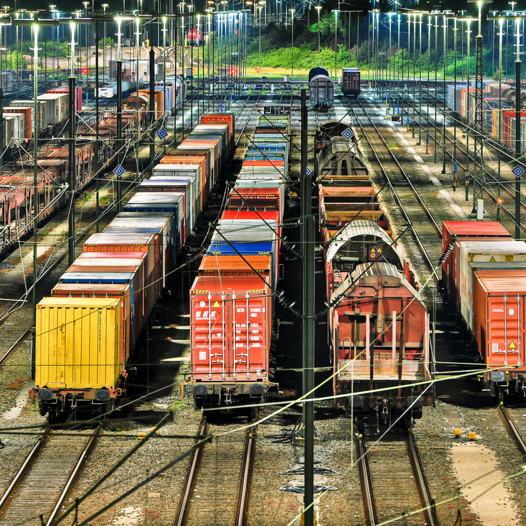 Ein nächtlicher Blick auf ein Güterbahnhofsgelände mit mehreren Gleisen, die von hellen Lampen beleuchtet werden. Zahlreiche Güterwaggons in verschiedenen Farben, darunter rote, gelbe und blaue Container, sind auf den Gleisen abgestellt. Im Hintergrund sind grüne Büsche und Bäume zu erkennen, die die Anlage begrenzen. Die Elektrifizierung ist deutlich erkennbar.