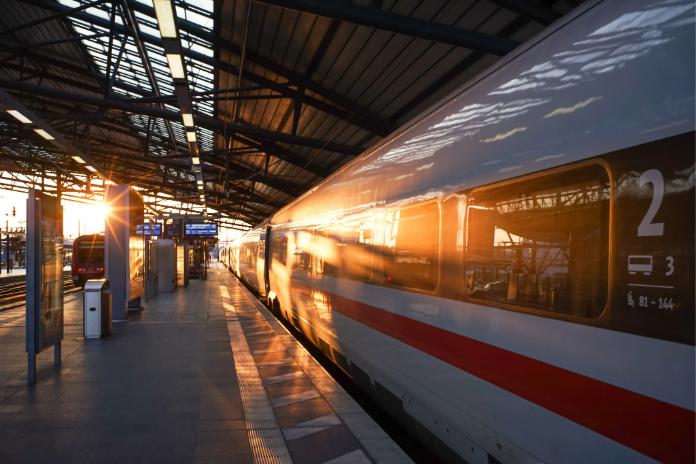 Ein weißer Hochgeschwindigkeitszug ICE mit roten Streifen steht an einem Bahnsteig, während die Sonne durch das Glasdach des Bahnhofs scheint und lange Schatten wirft. 