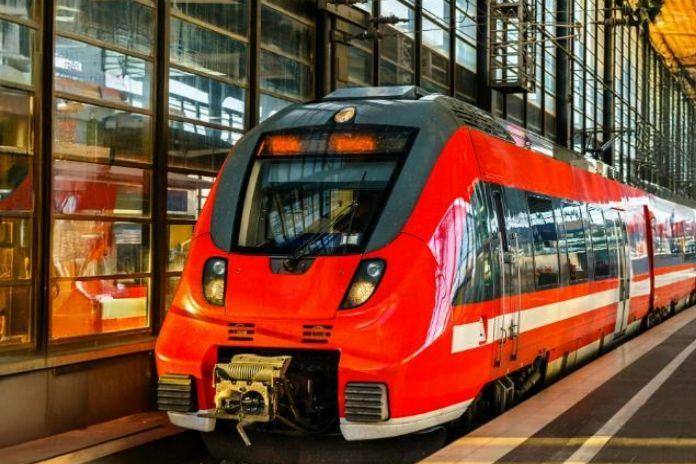 Ein moderner, roter Regionalzug Talent 2 mit weißen Streifen steht an einem Bahnsteig mit großen Glasfenstern. Der Zug scheint auf Fahrgäste zu warten.