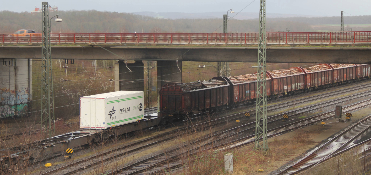 DLR-Forschungscontainer FR8-LAB auf einem Güterzug unter einer roten Brücke, im Hintergrund eine hügelige Landschaft.