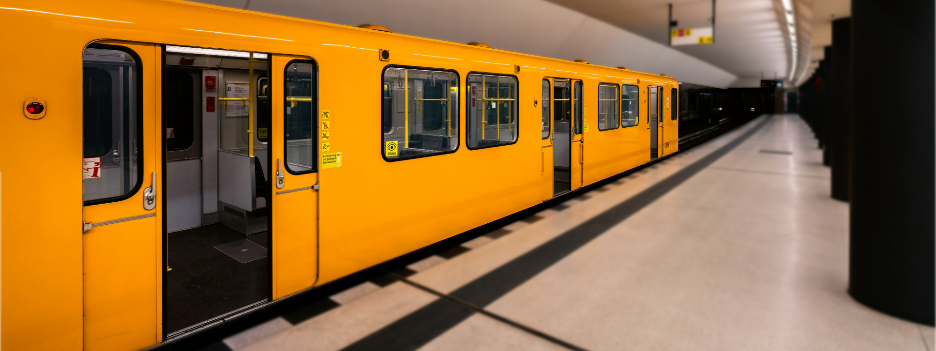 Ein leuchtend gelber U-Bahn-Zug der Berliner Verkehrsbetriebe (BVG) steht an einem Bahnsteig. Die Türen des Zuges sind geöffnet und geben den Blick auf den leeren Innenraum frei. Der Bahnsteig ist hell beleuchtet und sauber, mit einem schwarzen Sicherheitsstreifen am Bahnsteigrand. Im Hintergrund verläuft der Tunnel in die Dunkelheit.